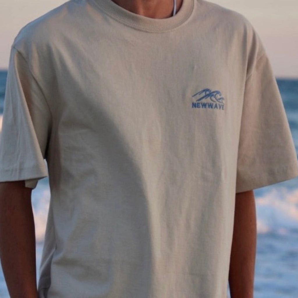 Person wearing a beige t-shirt with a blue logo and text, standing by the ocean.