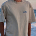 Person wearing a beige t-shirt with a blue logo and text, standing by the ocean.