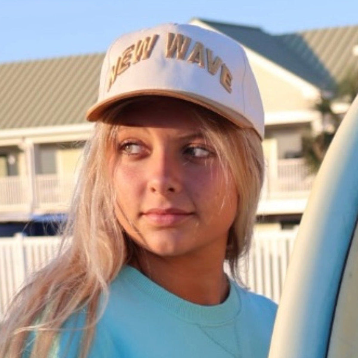 Person holding a surfboard with a logo, wearing a cap and light blue shirt, standing in front of a building.